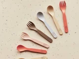 Seks farverige babyskeer og -sporker af plast med søde dyreansigter på skafterne er anbragt diagonalt på en plettet, beige overflade.