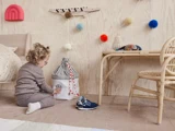 Et barn med krøllet hår, der har briller på og er klædt i behageligt tøj, leger på et trægulv ved siden af en stribet kurv i et pyntet rum med en rattan-stol, sko og farverige pomponer på væggen.