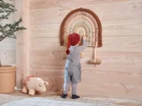 Et lille barn i overalls og en rød hue strækker sig op for at lege med et blødt aktivitetsbræt i form af en regnbue på væggen i et hyggeligt rum med træpaneler. Et udstoppet griselegetøj ligger lige v