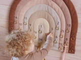 Et lille barn med krøllet hår rækker ud for at hænge en ring på en adventskalender af stof i form af en regnbue med nummererede lommer.