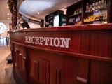 En receptionsskranke i træ med ordet RECEPTION i hvide bogstaver, placeret i et varmt oplyst interiør med hylder og en skranke i baggrunden.