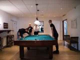 Fire unge mænd spiller pool i et veloplyst rum med trægulv; en tager et slag, mens de andre ser til rundt om bordet.