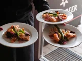 En person holder tre hvide tallerkener, hver med en gourmetret med laks, asparges, cherrytomater og syltede løg.