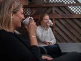 To kvinder sidder udendørs på en terrasse, smiler og drikker af hvide krus. Bag dem er der et træhegn, og i forgrunden står et bord.