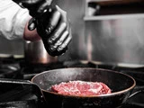 En person iført sorte handsker drysser salt på en rå bøf i en stegepande på komfuret.