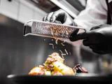 En kok iført sorte handsker river trøffel over en gourmetret på en tallerken i et professionelt køkken.