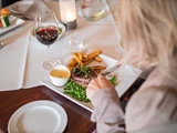 En person skærer i en bøf, der serveres med grønne ærter, pommes frites og sauce på en hvid tallerken, med et glas rødvin og et stearinlys på bordet.