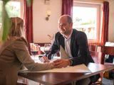 En mand og en kvinde sidder ved et restaurantbord, holder hinanden i hånden og smiler til hinanden med et glas rødvin foran sig.