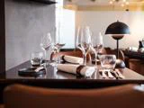 Et dækket spisebord i en restaurant med vinglas, vandglas, tallerkener, bestik, rullede servietter og en moderne sort bordlampe på en mørk, blank overflade.