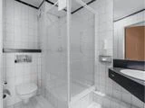 Moderne badeværelse med hvide flisevægge og -gulve, brusekabine i glas, væghængt toilet og sort bordplade med vask og spejl.
