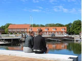 En mand og en kvinde sidder på en kaj ved vandet, med ryggen til hinanden, med bygninger med røde tage og grønne omgivelser i baggrunden under en blå himmel med spredte skyer.