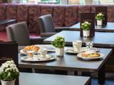Et cafébord dækket med kaffekopper, en croissant, et stykke kage, vandglas, hvide blomster i potter og menukort, med flere borde og stole i baggrunden.