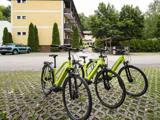 Tre grønne cykler er parkeret på en mønstret betonplads foran et gult lejlighedskompleks med træer og parkerede biler i nærheden.