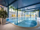 Indendørs swimmingpool med blåt loft, gule liggestole langs væggene, store vinduer, grønne planter og stærkt naturligt lys.