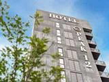 Høj moderne bygning med HOTEL PEAK 12 skrevet på facaden, fotograferet nedefra med en klar blå himmel og grønne trægrene i forgrunden.