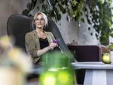En kvinde med briller sidder i en moderne stol med en lilla drink i hånden. Hun er klædt i en lys blazer og er omgivet af indendørs planter og hyggelig indretning.