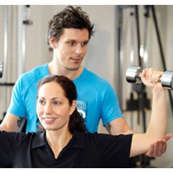 En kvinde løfter håndvægte, mens en mandlig træner i blå skjorte hjælper hende i fitnesscentret.
