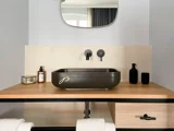 Moderne badeværelse med en rektangulær sort håndvask, vægmonteret vandhane, bordplade i træ, sæbedispensere, spejl og håndklæder nedenunder.