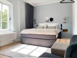 Moderne soveværelse med en stor seng, to puder, væglamper, et lille indrammet billede, et lyst tæppe, et vindue med sollys, en radiator og minimalistisk indretning.