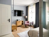 Lyst, moderne soveværelse med et vægmonteret tv, et træskab, en lilla accentstol, en gulvlampe, store vinduer med gardiner og en seng, der delvist er synlig.