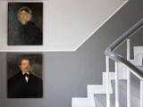 To indrammede portrætter hænger på en grå væg ved siden af en hvid og grå trappe med et gelænder af metal.
