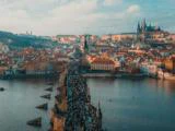 Luftfoto af den overfyldte Karlsbro, der spænder over Moldau, med historiske bygninger og Prags borg i baggrunden under en delvis overskyet himmel.