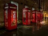 En række klassiske røde britiske telefonbokse er opstillet langs en våd, svagt oplyst gade ved siden af en murstensbygning om natten.