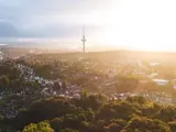 Luftfoto af en by med spredte huse, tætte grønne træer og et højt kommunikationstårn under en lys, diset himmel ved solopgang eller solnedgang.