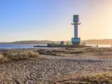 Et fyrtårn står på en mole ved vandet med sandstrand og tørt græs i forgrunden under en klar blå himmel ved solnedgang.