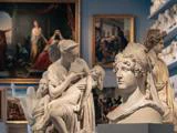 Klassiske skulpturer og buster udstillet i et museumsgalleri med indrammede malerier på væggene i baggrunden.
