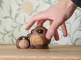 En hånd rækker ud mod to små, runde fuglefigurer af træ med store, sorte øjne, som sidder på en træplade mod en tapetbaggrund med blade.