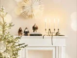 En kaminhylde pyntet med tre små julemandsfigurer, bøger, en moderne lysestage til fire stearinlys og en fyrretræsgren. I baggrunden ses et tændt juletræ og en vægdekoration i form af en stjerne.