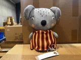 Plys-koala med et gråt mønstret hoved og en brun og orange stribet kjole, med en papirmærkat fastgjort.