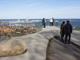Flere mennesker står på et moderne observationsdæk af glas med udsigt over en kystby, havnen og havet under en delvis overskyet himmel. En hund er også til stede.
