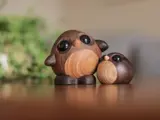 To små, runde uglefigurer af træ med store, sorte øjne er placeret på en glat overflade med en sløret, grøn plante i baggrunden.