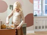 Et lille barn med lyst hår leger med et legetøjskøkken og bruger en ske til at røre i mad. Værelset er indrettet i pastelfarver og har et stort vindue, der lukker naturligt lys ind.