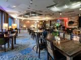 Rummelig, moderne restaurant med træborde og -stole, dekorativ belysning, mønstret gulvtæppe og planter på bordene. Store vinduer og varm belysning skaber en hyggelig atmosfære.