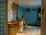 To personer i et spa-rum med blå flisebelagte vægge; den ene sidder ved et træbadekar, den anden står under en bruser iført stribede shorts.