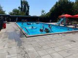 Børn og voksne svømmer og leger med svømmehjælpemidler i en udendørs pool, hvor der er en rutsjebane og parasoller i nærheden på en solskinsdag.