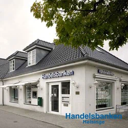 En hvid murstensbygning med tegltag huser Handelsbankens filial i Helsinge. Store vinduer viser plakater, og en grøn postkasse er monteret nær indgangen.