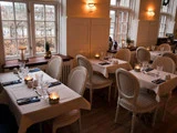 Elegant restaurant med hvide duge, stearinlys, glas og vintagestole ved siden af store vinduer, der lukker naturligt lys ind.