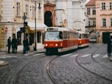 En rød og hvid sporvogn kører ad buede skinner på en brostensbelagt gade i en europæisk by, hvor den passerer gamle bygninger og fodgængere.