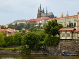 En udsigt over Prags borg og de omkringliggende historiske bygninger på en bakke med træer og en flod i forgrunden under en overskyet himmel.