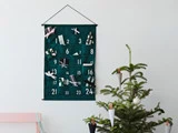 En grøn stofadventskalender med lommer nummereret 1-24 hænger på en væg over et pyntet juletræ med koglepynt.