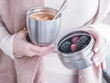 En person med en rustfri stålbeholder med bær i den ene hånd og en kop kaffe med en ske i den anden, iført en lys sweater og vest.