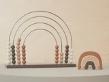 En regnbueformet kugleramme med neutralfarvede perler ved siden af en lille regnbuestabler i træ i lignende jordfarver, begge på en lysegrå overflade.