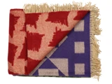 Et foldet uldtæppe med beige frynser, der viser et rødt og beige hundetandsmønster på toppen og et lilla og beige geometrisk mønster på det bageste hjørne.