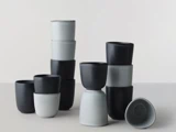 En gruppe minimalistiske keramiske kopper i sort og lysegrå, stablet i forskellige arrangementer på en neutral overflade.