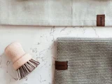 En skrubbebørste af træ, en struktureret grå klud og et beige stof med brune mærker arrangeret på en hvid marmoroverflade.
