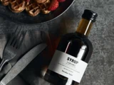 En flaske karamelsirup ligger ved siden af en gaffel og en kniv på en serviet, og en tallerken med vafler og jordbær er delvist synlig ovenover.